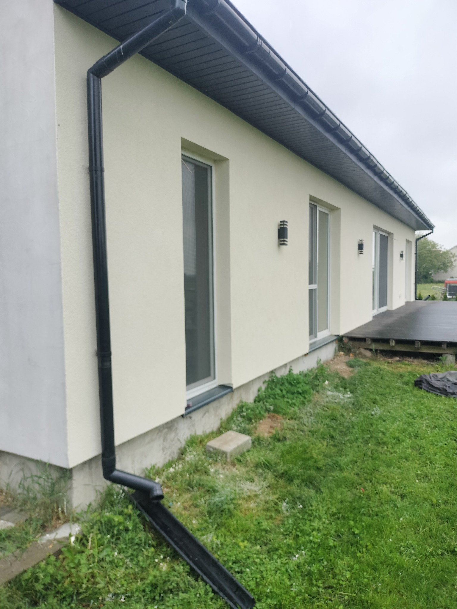 Elewacja domu jednorodzinnego z zamontowanym systemem rynnowym w kolorze grafitowym. Widoczne okna i lampy zewnętrzne. Trawa i fragment tarasu.