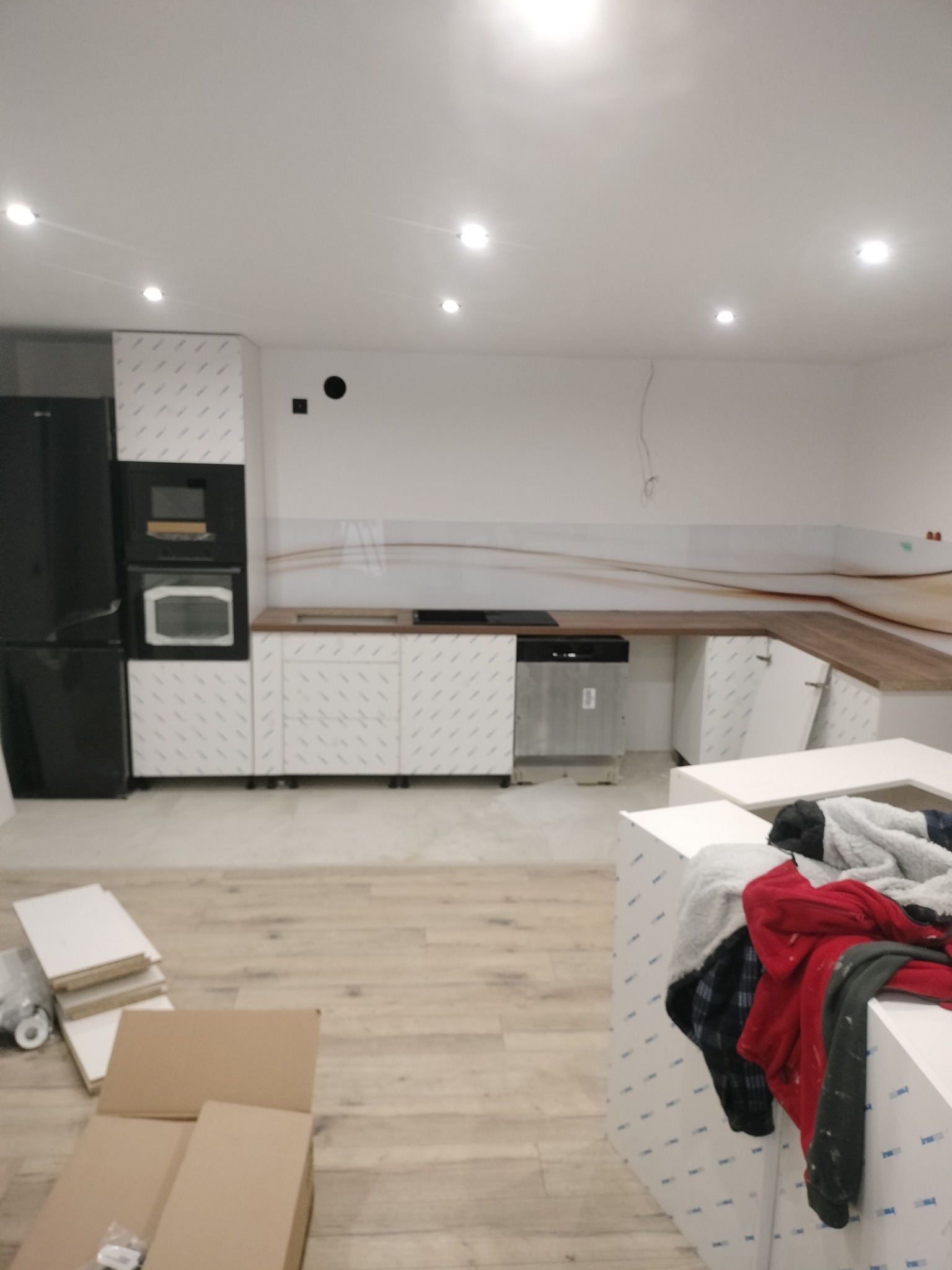 Wnętrze kuchni w trakcie montażu: białe szafki z folią ochronną, drewniany blat, sprzęt AGD. Na podłodze kartony i płyty, ubrania na wyspie kuchennej.
