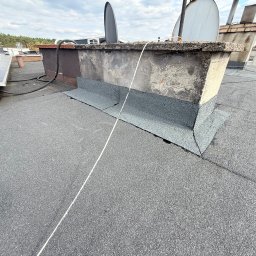 HydroDach System - Szary dach pokryty papą z wykończonym kominem i panelami słonecznymi w tle. Widoczne detale łączeń i faktura materiału. Ujęcie z góry, podkreślające solidność wykonania.