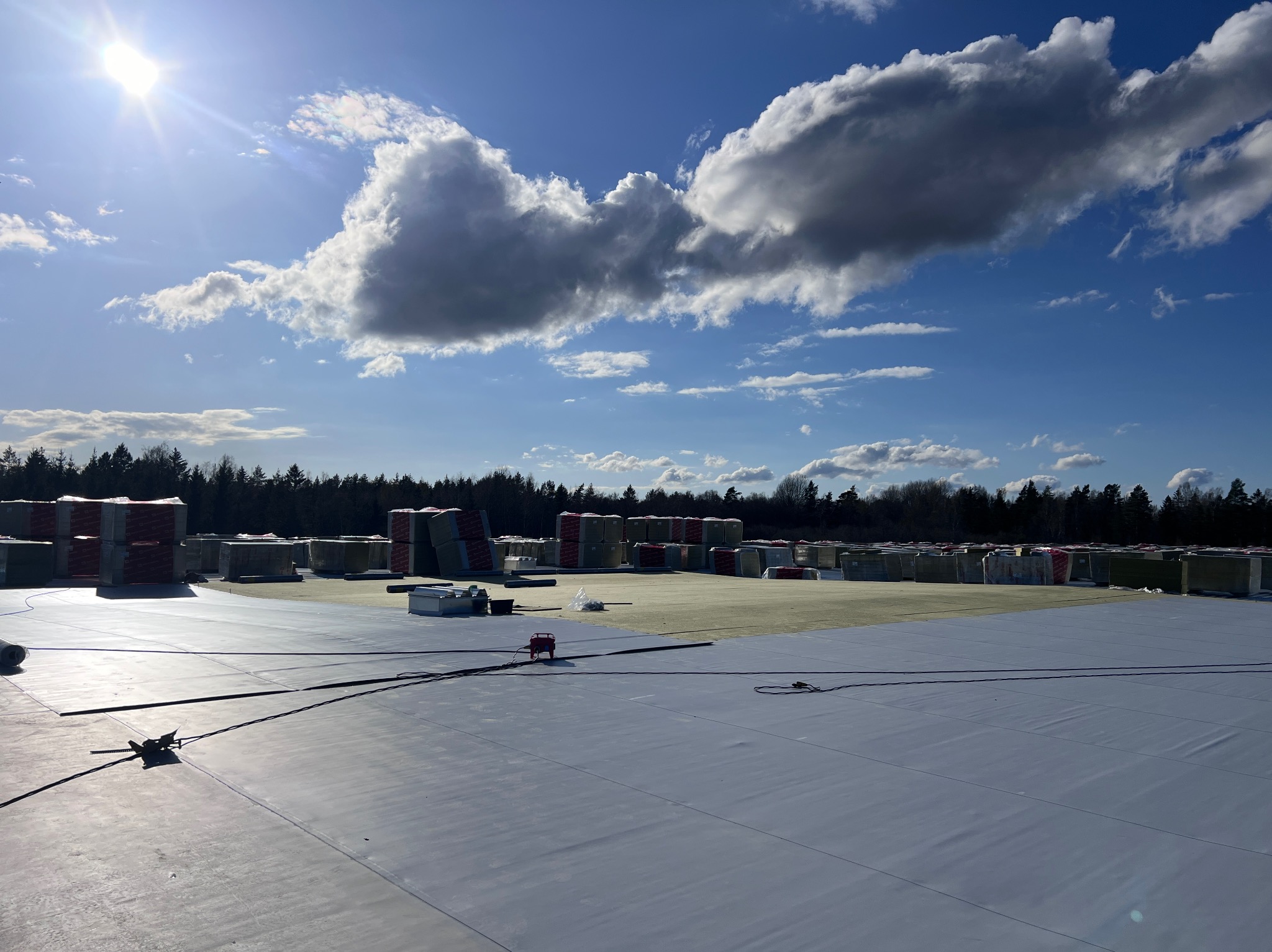 Rozległy, płaski dach pokryty szarą membraną, z przygotowanymi do montażu pakietami izolacyjnymi w tle, pod błękitnym niebem z chmurami i słońcem.