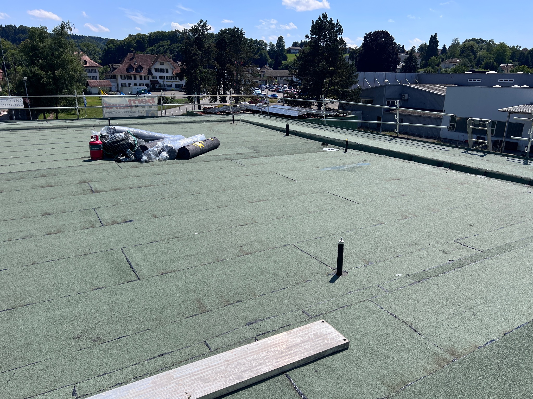 Zielony dach płaski z papy termozgrzewalnej w trakcie prac. Widoczne rolki papy, butla gazowa i narzędzia. W tle zabudowa miejska i zieleń.