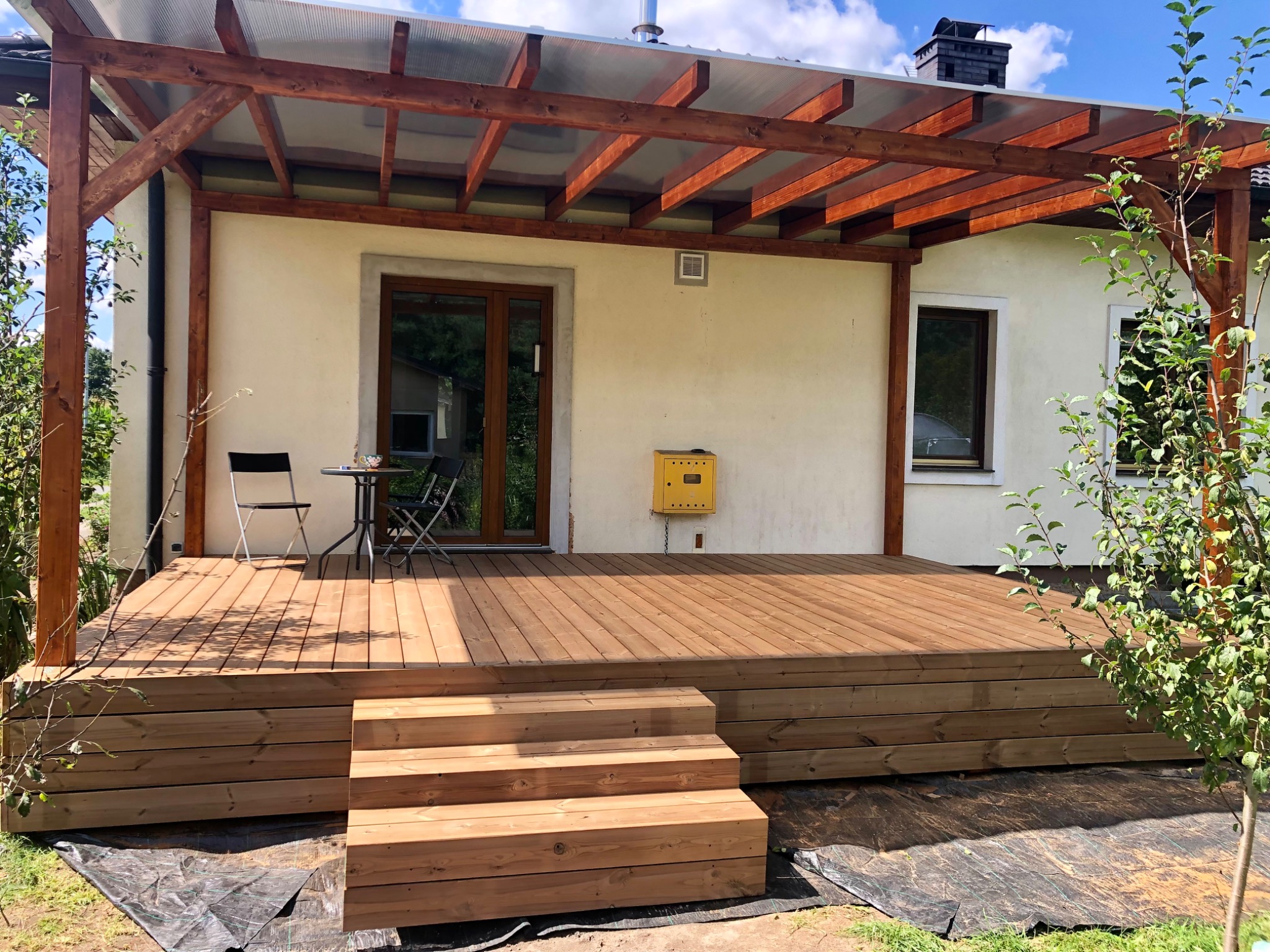 Zadaszony taras z drewna z niskimi schodami, stolikiem i krzesłami. Widoczna elewacja domu z oknami i żółtą skrzynką. Całość w słoneczny dzień.