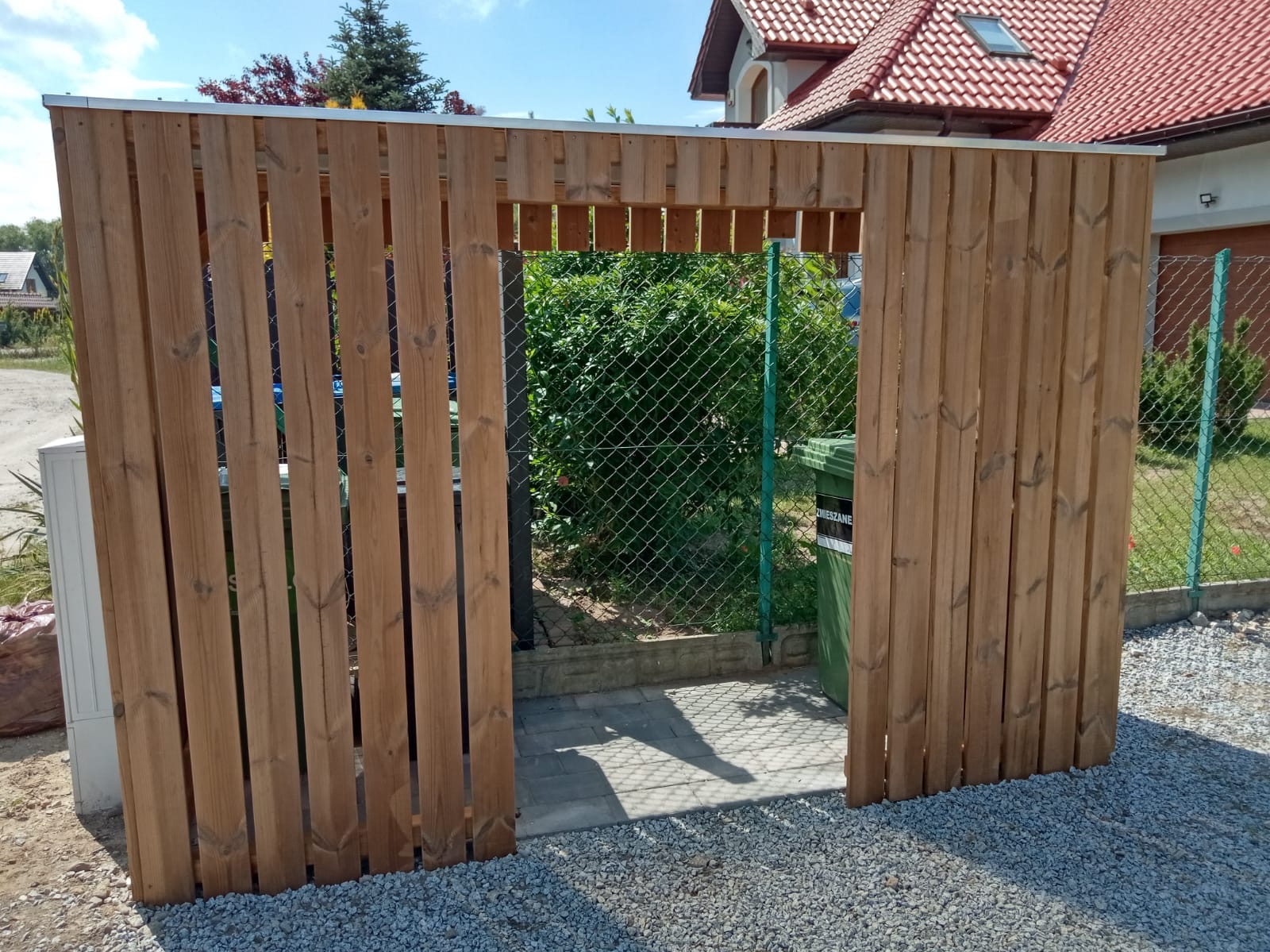 Drewniana osłona śmietnika z pionowych desek, z wejściem, na tle zieleni i domu z czerwoną dachówką, na żwirowym podłożu. Widoczny kosz na śmieci.