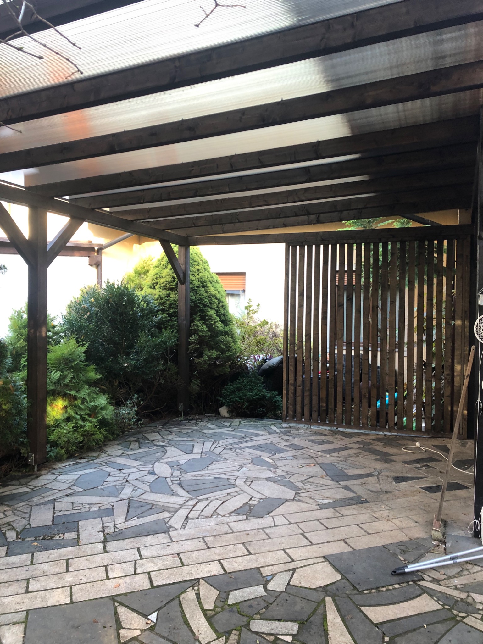 Zadaszona konstrukcja z ciemnego drewna, prawdopodobnie pergola, z widocznym ażurowym ogrodzeniem i nieregularną kamienną nawierzchnią. Zieleń w tle.