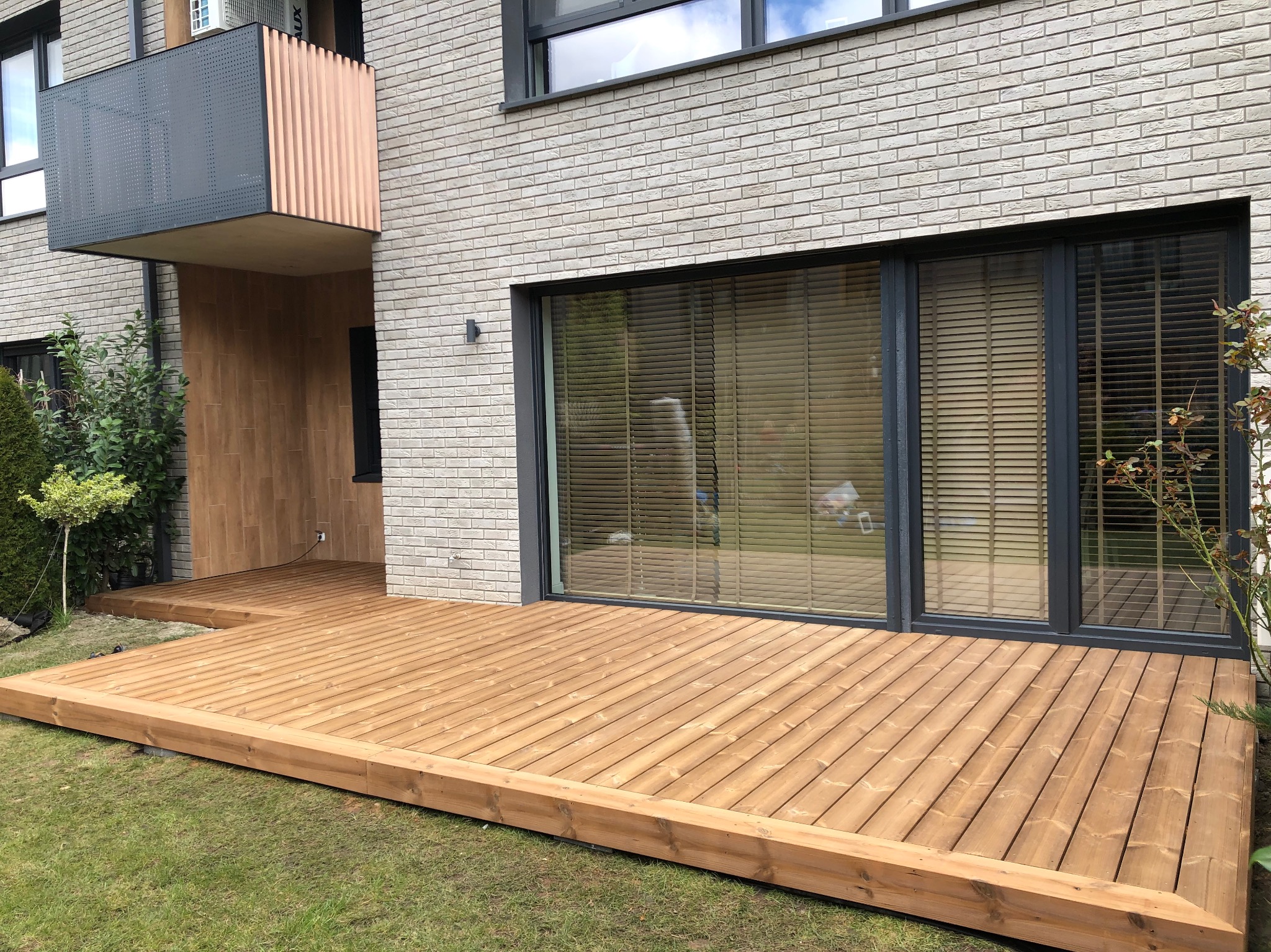 Jasny taras drewniany przylegający do nowoczesnego domu z cegły. Widoczne duże okno przesuwne i balkon z metalowymi elementami. Trawa wokół tarasu.
