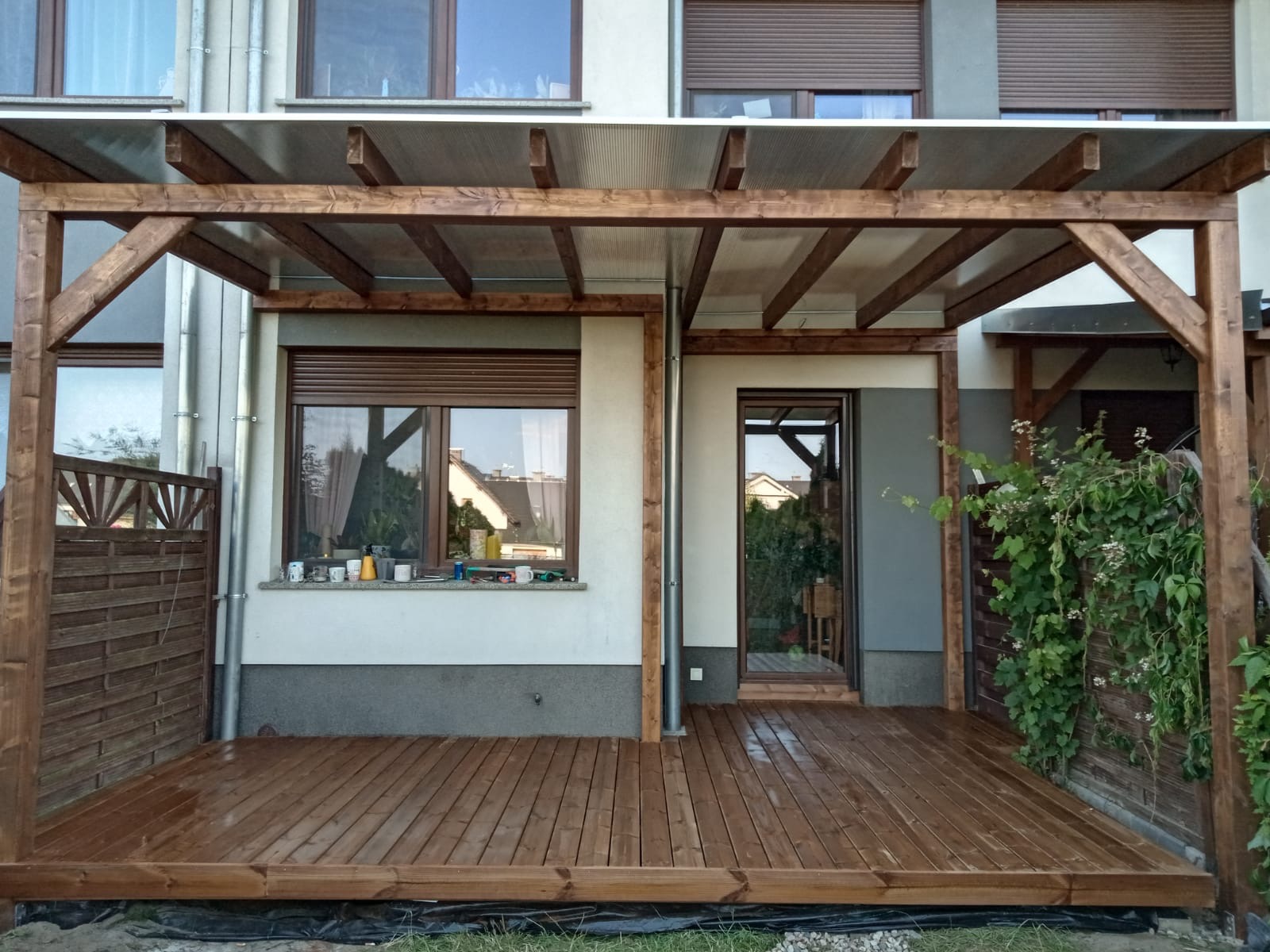 Zadaszony taras z drewna w odcieniach brązu, przylegający do budynku z oknami i drzwiami. Widoczne elementy konstrukcyjne i deski tarasowe. Na parapecie okna stoją kubki.