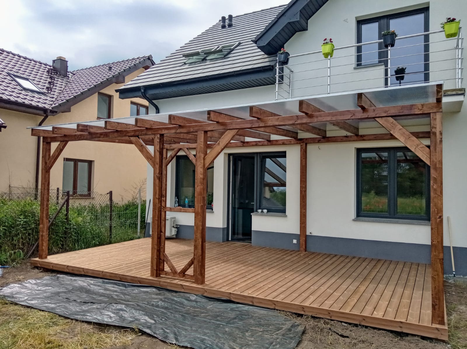 Drewniana pergola z przezroczystym zadaszeniem, połączona z tarasem z desek. Widok na dom z balkonem i sąsiedni budynek. Ochronna plandeka na ziemi.