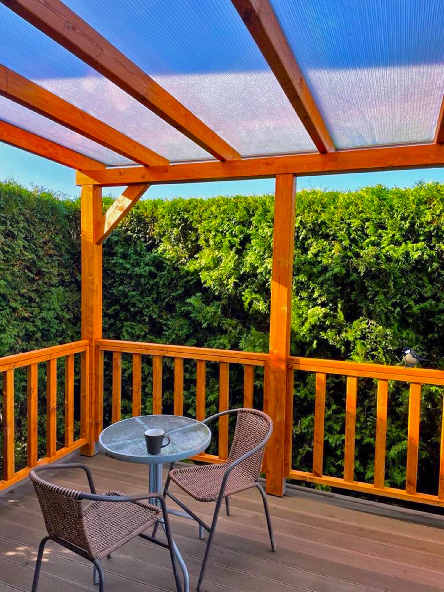 Zadaszony taras z drewnianą konstrukcją i przezroczystym dachem. Na tarasie stolik z filiżanką i dwa krzesła. W tle zielony żywopłot i błękitne niebo.