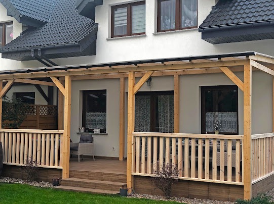 Zadaszony taras z drewnianą konstrukcją i balustradą, przylegający do domu. Widoczne meble ogrodowe i schody wejściowe. Jasne drewno, schludny wygląd.