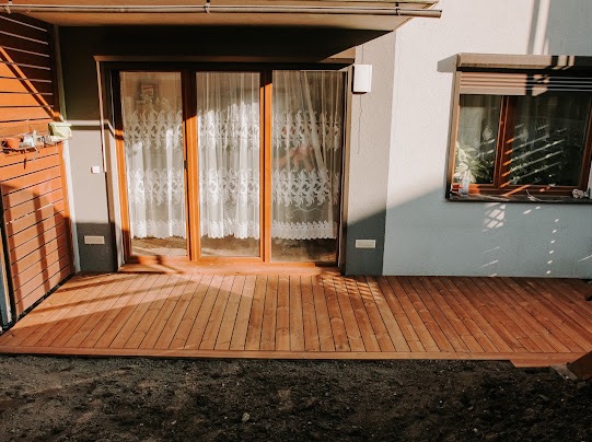 Nowoczesny taras z desek w ciepłym odcieniu brązu, przylegający do domu z dużymi oknami zasłoniętymi koronkowymi firankami. Ujęcie w słoneczny dzień.