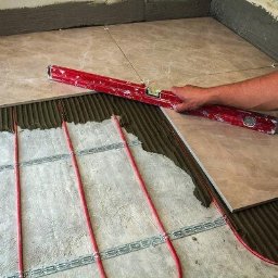 Układanie płytek na ogrzewaniu podłogowym. Czerwone rurki, poziomica w dłoni, beżowe płytki z marmurkowym wzorem. Widoczny klej i fragment ściany.