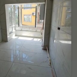 Jasna łazienka z białymi, marmurkowymi płytkami podłogowymi i ściennymi. Widoczne okno z widokiem na żółty budynek. Instalacja elektryczna i rury wzdłuż ściany.