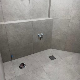 FAYANS DOMINIKA ARAT - Wykończona kabina prysznicowa z szarymi, matowymi płytkami. Widoczne odpływy i przyłącza wodne. Minimalistyczny design, precyzyjne fugi, nowoczesne rozwiązanie.