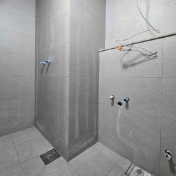 FAYANS DOMINIKA ARAT - Wykończona, szara łazienka z odpływem liniowym i wystającymi rurami. Minimalistyczny design, płytki imitujące kamień, surowy styl. W tle wieszak na ubrania.