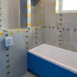 FAYANS DOMINIKA ARAT - Łazienka w trakcie wykańczania: jasne płytki imitujące marmur, system poziomowania płytek z żółtymi klinami, niebieska obudowa wanny, biała armatura i okno.