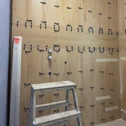 FAYANS DOMINIKA ARAT - Ściana w trakcie wykańczania drewnopodobnymi płytkami, z użyciem systemu poziomowania. Widoczna drabina i fragment podłogi. Oświetlenie sufitowe.