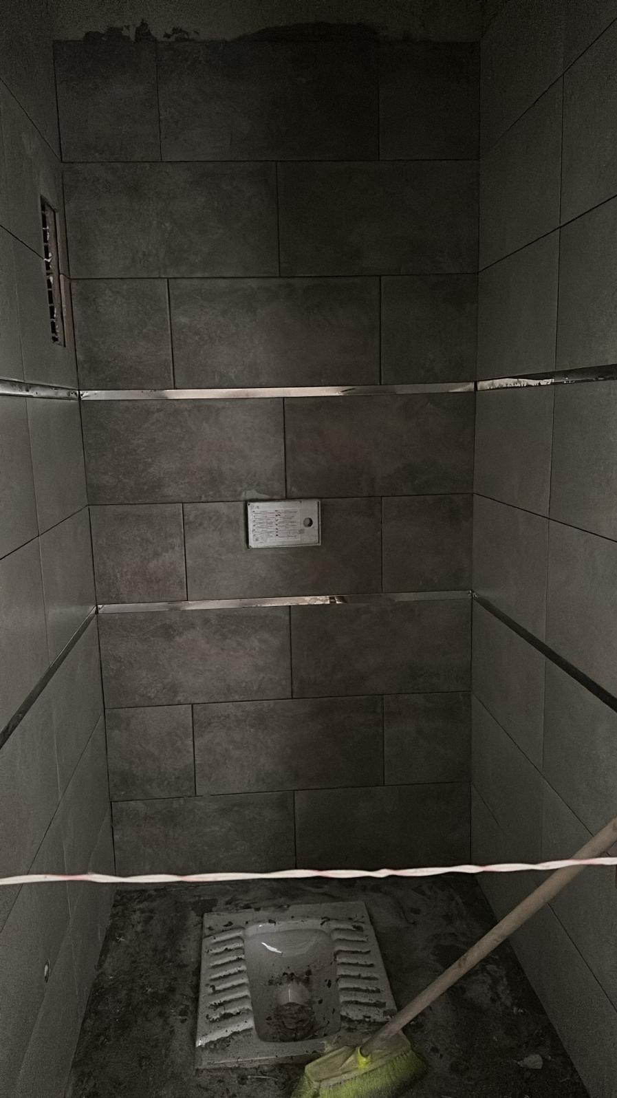 Szara łazienka w trakcie remontu. Widoczne płytki na ścianach z metalowymi listwami, stara toaleta i miotła. Surowy stan wykończenia.