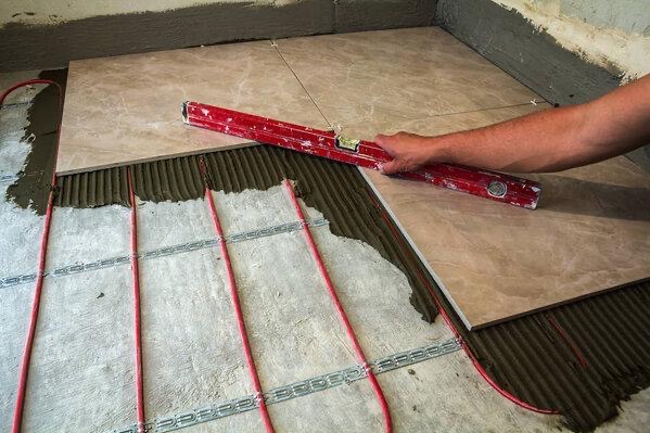Układanie płytek na ogrzewaniu podłogowym. Czerwone rurki, poziomica w dłoni, beżowe płytki z marmurkowym wzorem. Widoczny klej i fragment ściany.