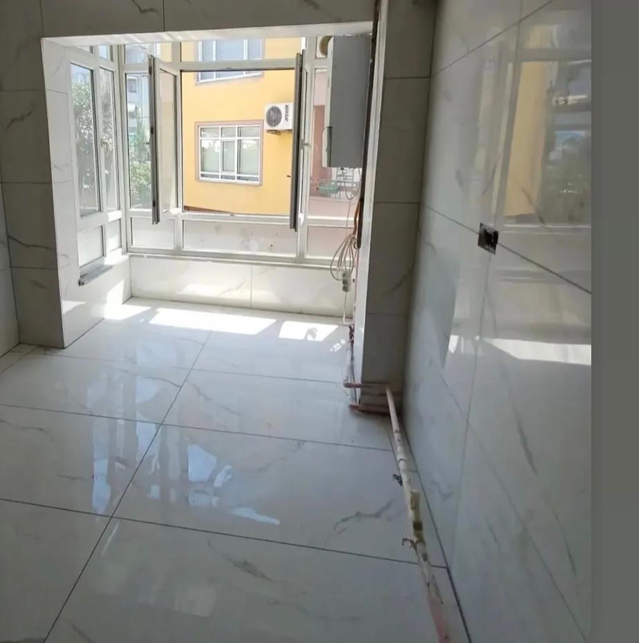 Jasna łazienka z białymi, marmurkowymi płytkami podłogowymi i ściennymi. Widoczne okno z widokiem na żółty budynek. Instalacja elektryczna i rury wzdłuż ściany.