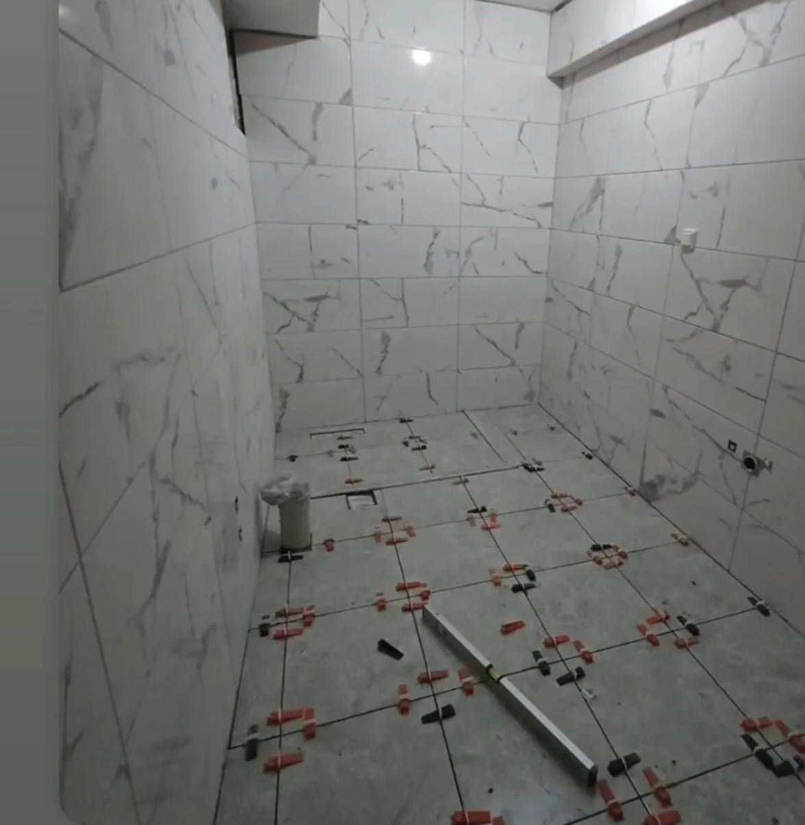 Białe płytki imitujące marmur na ścianach i podłodze łazienki w trakcie układania. Widoczne krzyżyki dystansowe i poziomica. Częściowo ułożona podłoga.