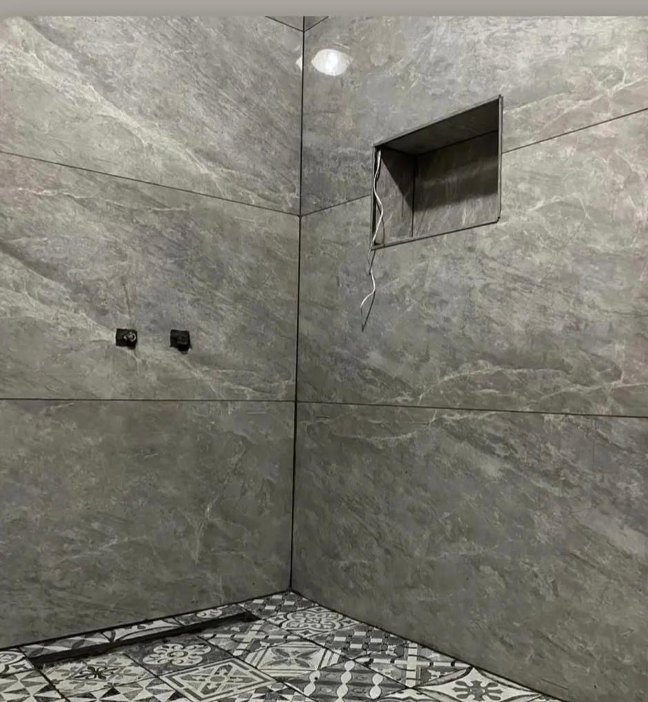 Wykończona łazienka z szarymi płytkami imitującymi marmur i dekoracyjną podłogą. Widoczna wnęka w ścianie z wystającymi kablami i gniazdka elektryczne.