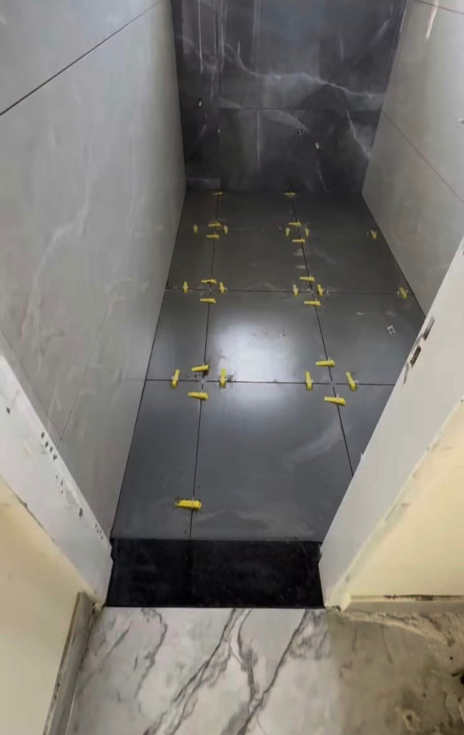 Układanie ciemnych płytek w łazience z użyciem żółtych krzyżyków dystansowych. Widoczne marmurowe wykończenia ścian i podłogi. Prace w toku, świeżo położone płytki.