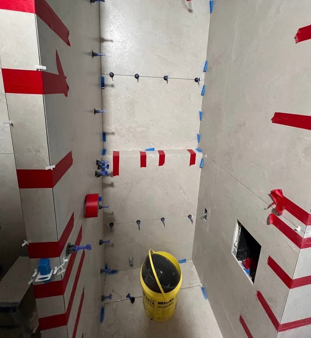 Świeżo ułożone, jasne płytki w łazience, zabezpieczone czerwoną taśmą i niebieskimi krzyżykami dystansowymi. Żółte wiadro na podłodze. Widok na fragment instalacji.