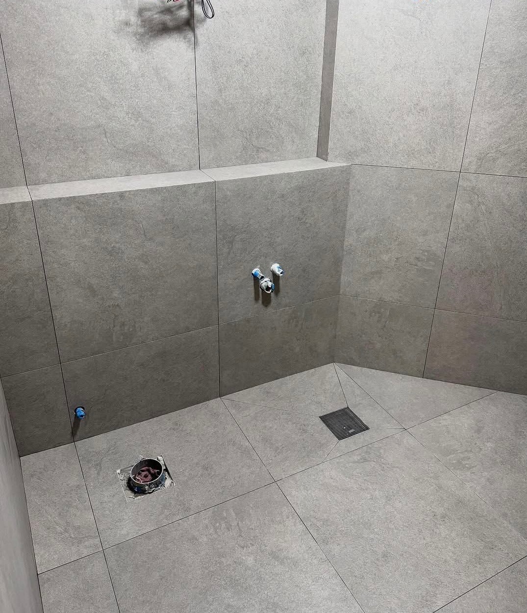 Wykończona kabina prysznicowa z szarymi, matowymi płytkami. Widoczne odpływy i przyłącza wodne. Minimalistyczny design, precyzyjne fugi, nowoczesne rozwiązanie.