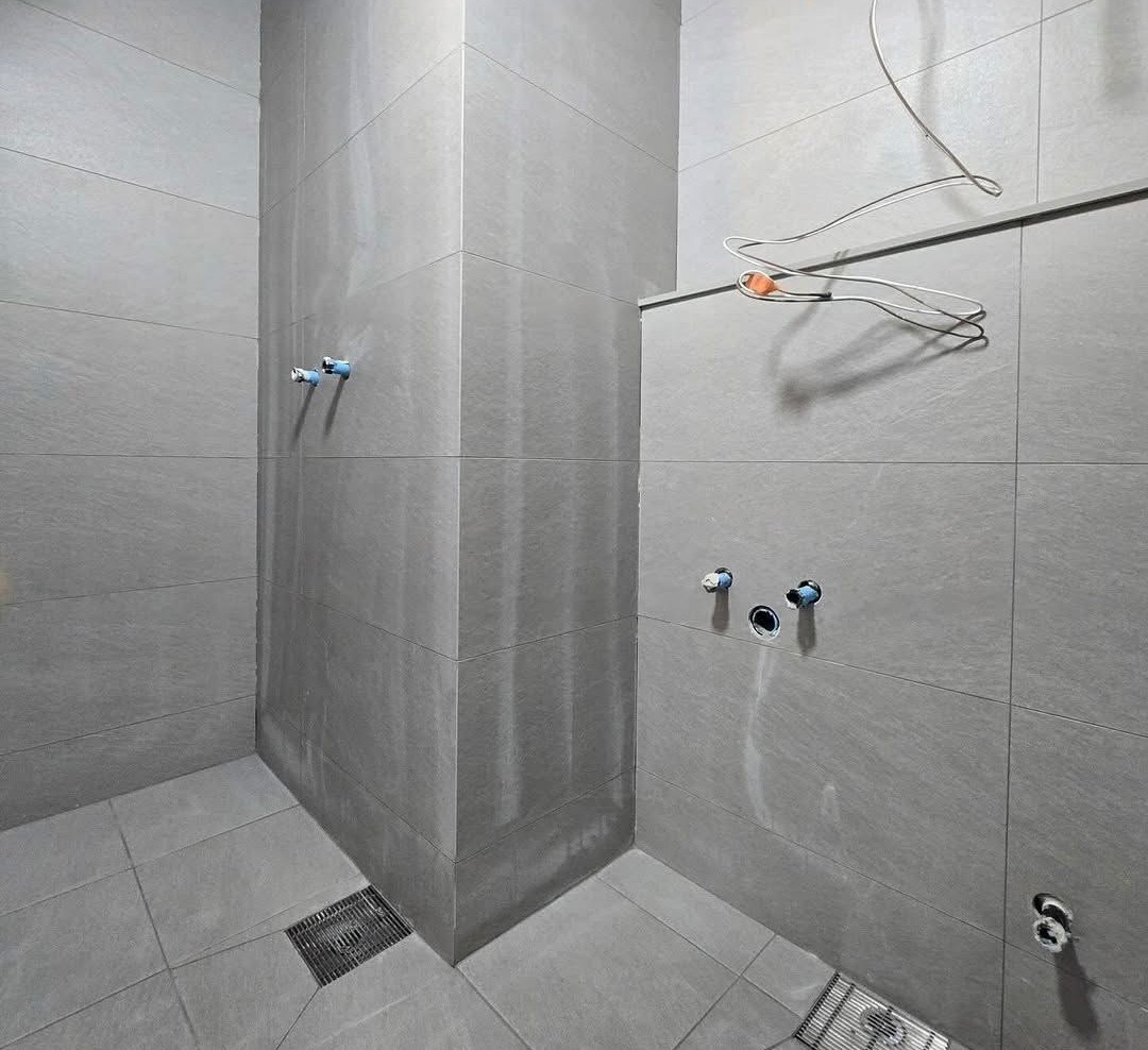 Wykończona, szara łazienka z odpływem liniowym i wystającymi rurami. Minimalistyczny design, płytki imitujące kamień, surowy styl. W tle wieszak na ubrania.