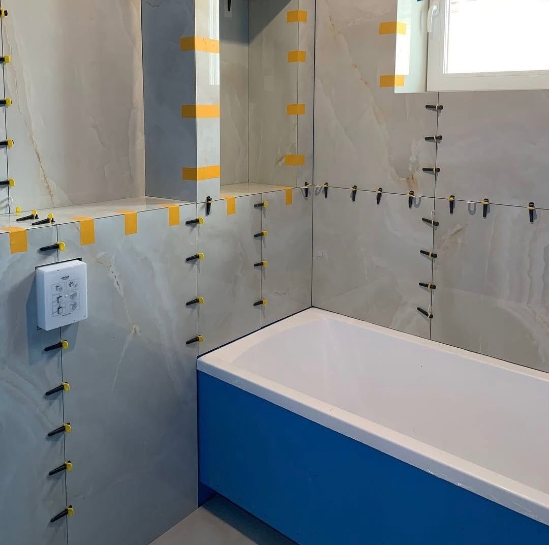 Łazienka w trakcie wykańczania: jasne płytki imitujące marmur, system poziomowania płytek z żółtymi klinami, niebieska obudowa wanny, biała armatura i okno.