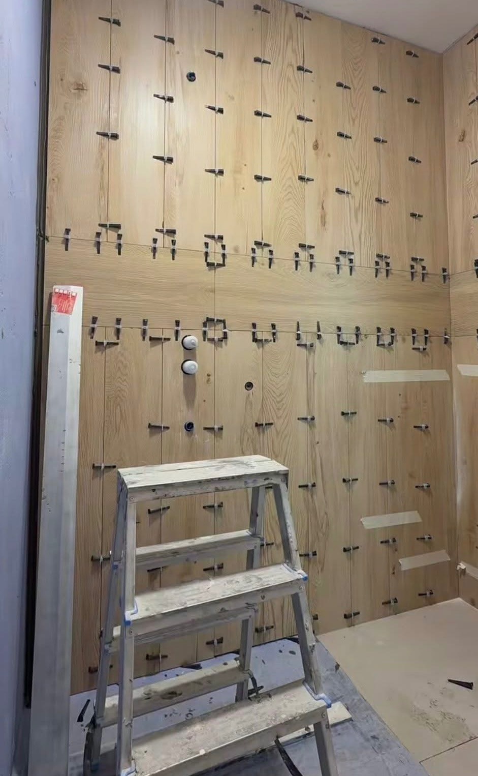 Ściana w trakcie wykańczania drewnopodobnymi płytkami, z użyciem systemu poziomowania. Widoczna drabina i fragment podłogi. Oświetlenie sufitowe.