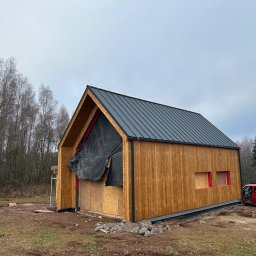 Usługi Ogólnobudowlane Jawor Mateusz Jaworski - Wznoszony dom szkieletowy z drewnianą elewacją i czarnym dachem na tle lasu. Część ściany zakryta folią. Czerwony bus zaparkowany obok.