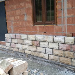 Wznoszenie muru z kamienia elewacyjnego na budynku z czerwonej cegły. Widoczne okno z brązową ramą i czarne drzwi. Częściowo wykonana elewacja z kamienia.