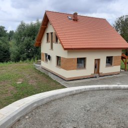 Usługi Ogólnobudowlane Jawor Mateusz Jaworski - Ukończony dom z beżową elewacją i czerwoną dachówką, widok z zewnątrz, z elementami wykończenia imitującymi drewno. Widoczny fragment podjazdu z kostki.