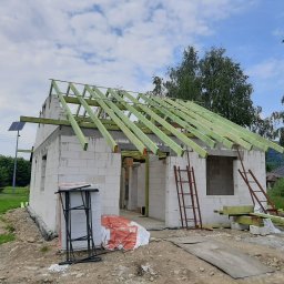 Usługi Ogólnobudowlane Jawor Mateusz Jaworski - Budowa domu z białych bloczków: ściany wzniesione, zielone krokwie dachu, drabiny i materiały budowlane na placu. Widok na budynek w trakcie realizacji.