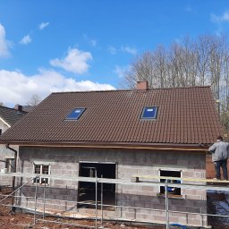 Usługi Ogólnobudowlane Jawor Mateusz Jaworski - Budowa domu z bloczków: surowy stan z brązowym dachem i oknami dachowymi. Na rusztowaniu stoi pracownik. W tle drugi budynek i drzewa.