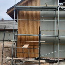 Usługi Ogólnobudowlane Jawor Mateusz Jaworski - Budowa domu szkieletowego: fragment elewacji z drewnianą boazerią i szarym tynkiem, widoczne rusztowania. Etap prac wykończeniowych.