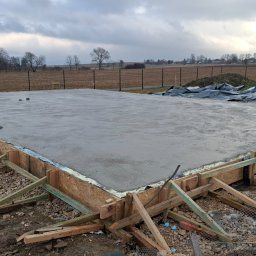 Zalany betonem fundament z drewnianym szalunkiem, widoczne niebieskie rury i otaczający teren z resztkami śniegu. Pochmurne niebo w tle.