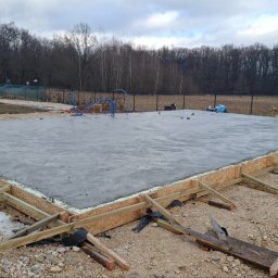 Zalana płyta fundamentowa z drewnianym szalunkiem na żwirowym podłożu. W tle drzewa i częściowo widoczny czerwony samochód dostawczy.