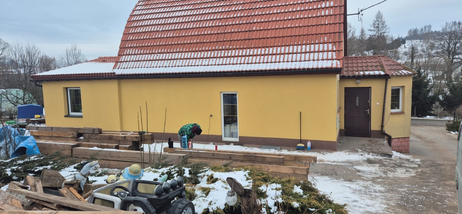 Żółty dom z czerwoną dachówką, częściowo pokryty śniegiem. W tle zalesione wzgórza. Na pierwszym planie drewniane elementy i osoba w trakcie pracy na zewnątrz.