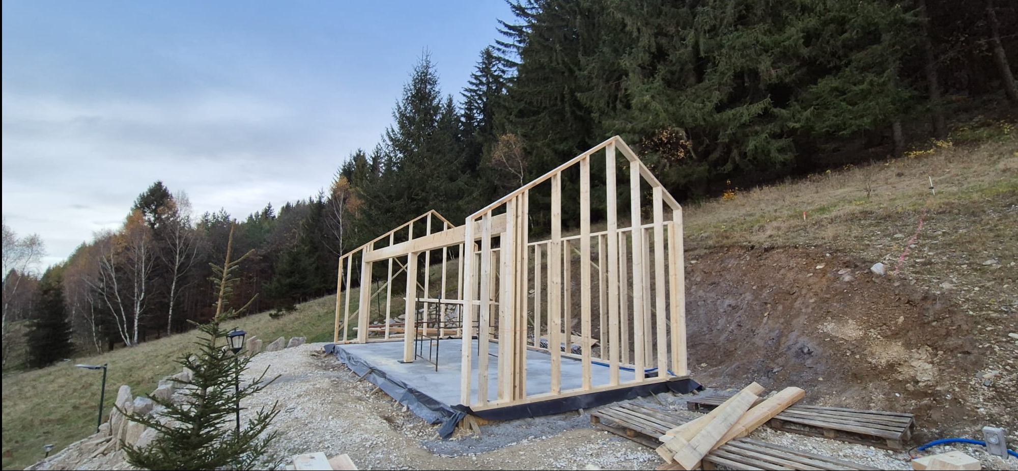 Wznoszenie szkieletu domu na betonowej płycie fundamentowej, widok na drewnianą konstrukcję na tle zalesionego wzgórza i pochmurnego nieba.