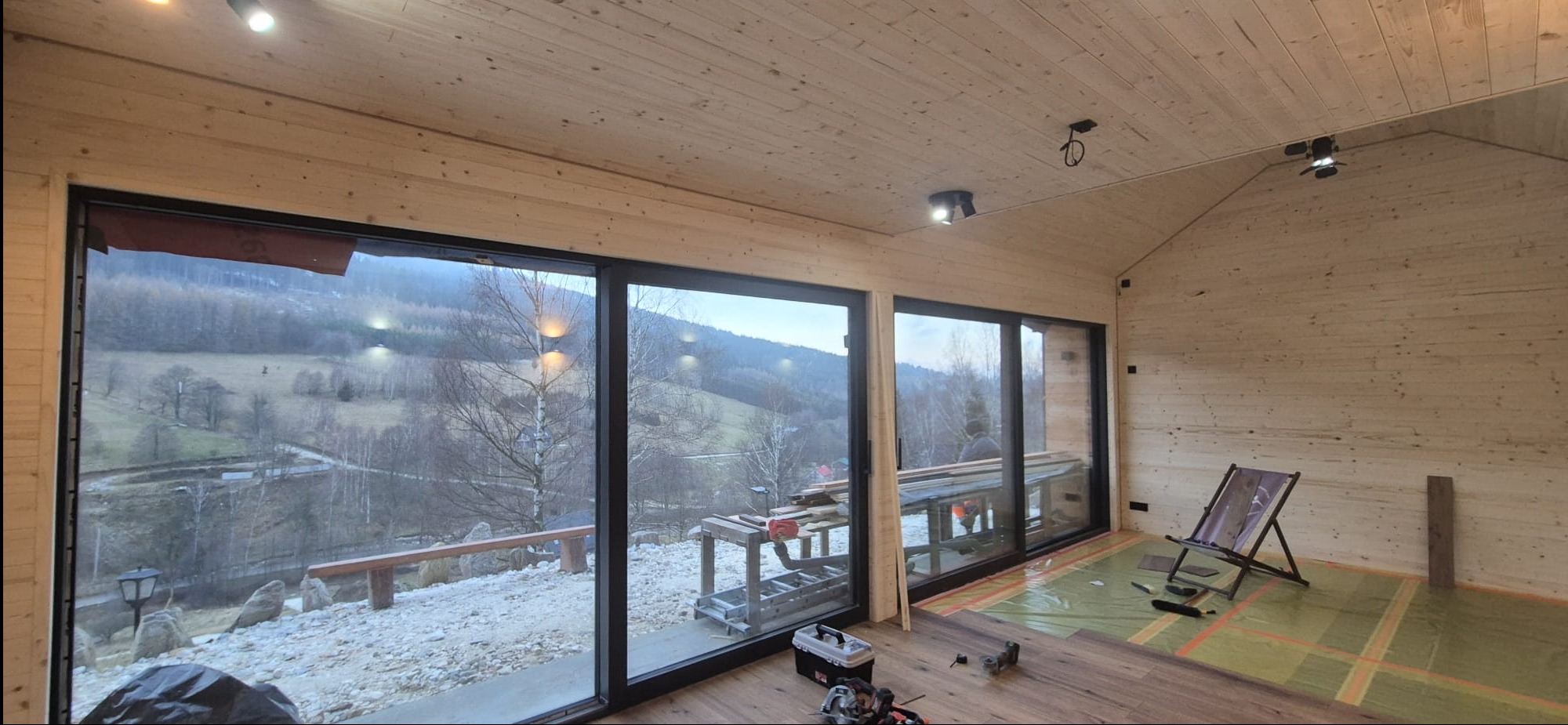 Wnętrze domu w trakcie wykańczania: drewniane ściany i sufit, duże okna z widokiem na góry, narzędzia i materiały do układania podłogi. Widoczny etap prac wykończeniowych.