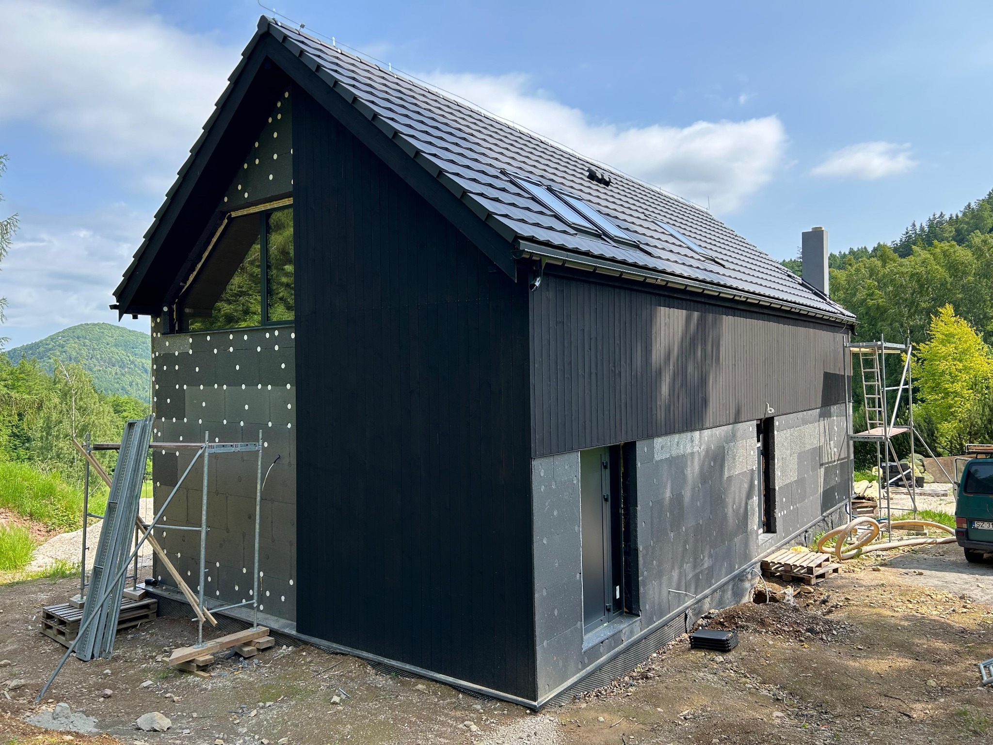 Nowoczesny dom w trakcie ocieplania elewacji, widoczne rusztowania i materiały izolacyjne. Część ściany wykończona czarnym drewnem, okna dachowe.