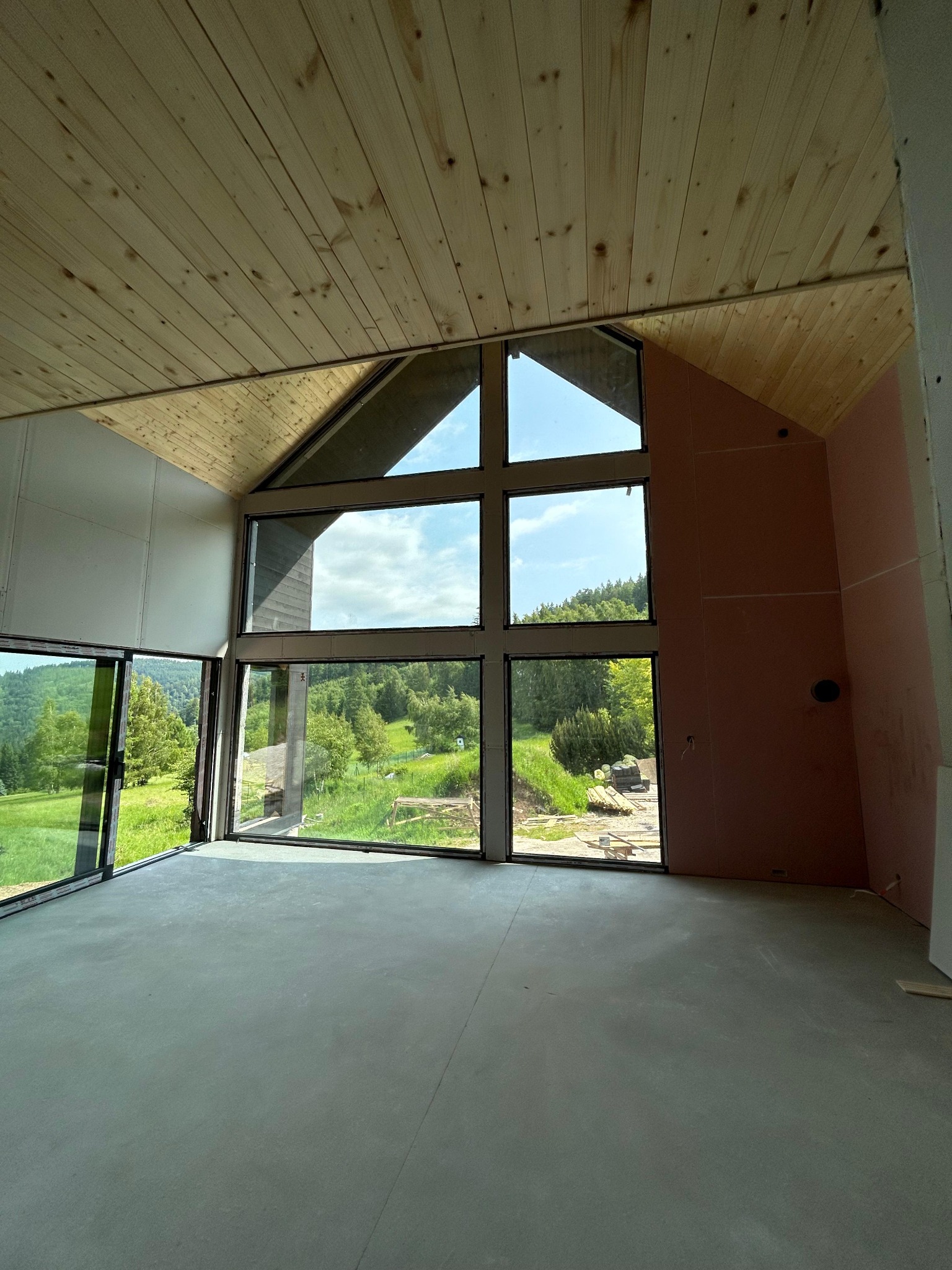 Wnętrze domu w trakcie budowy z dużymi oknami z widokiem na zieleń. Ściany częściowo wykończone płytami, drewniany sufit. Widoczny krajobraz za oknem.