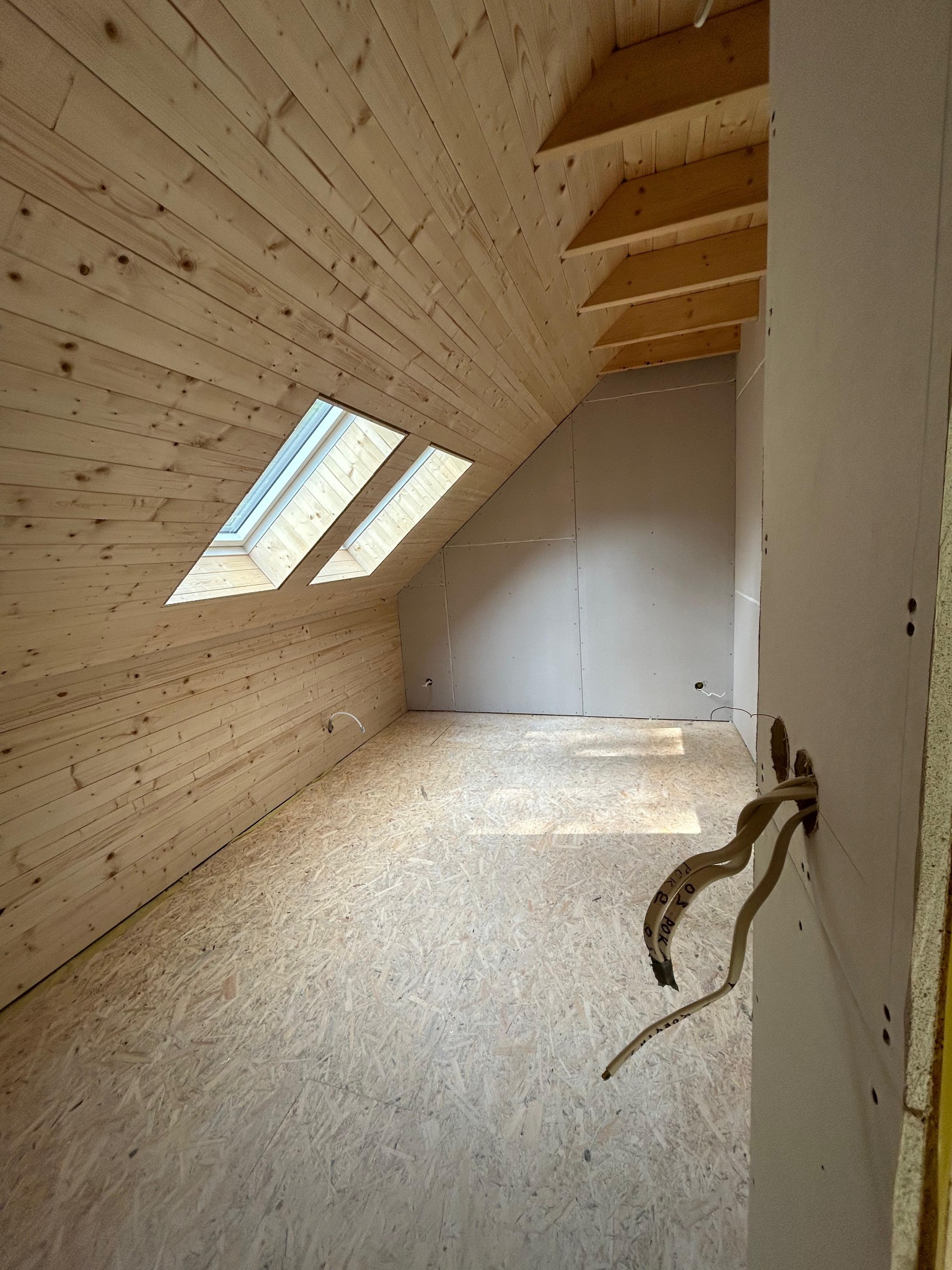 Poddasze z drewnianą boazerią, dwoma oknami dachowymi i płytami kartonowo-gipsowymi na ścianach. Widoczne wystające kable elektryczne.