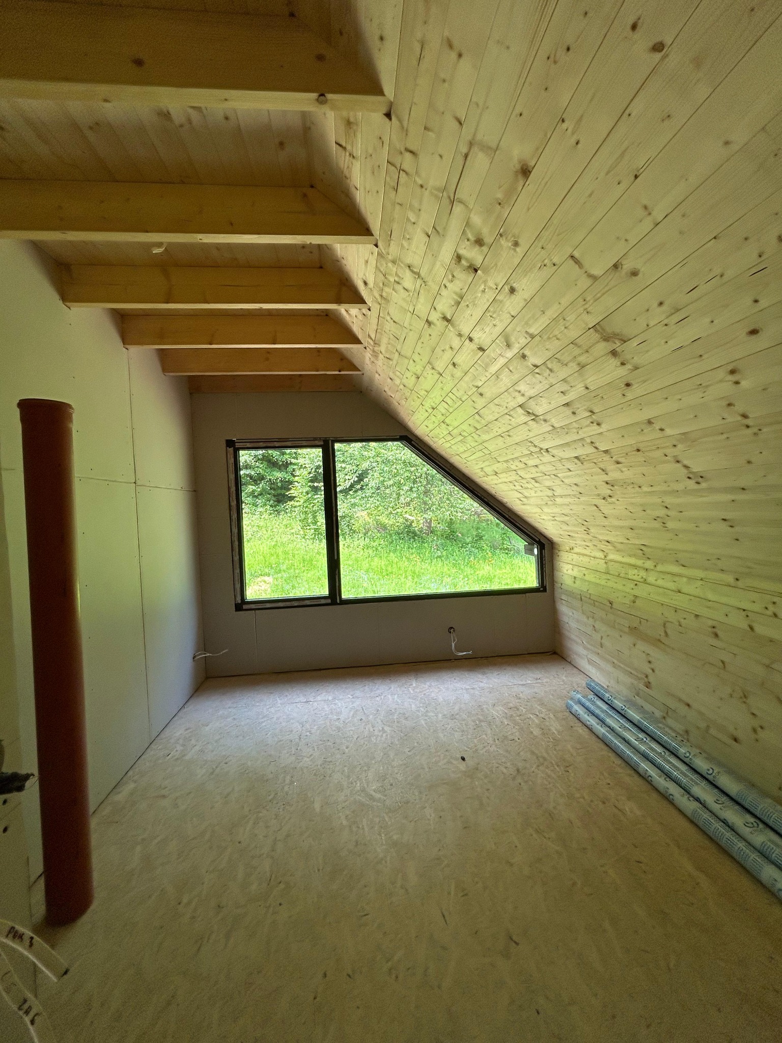 Poddasze z oknem w kształcie trapezu, wykończone boazerią i płytami gipsowymi, z widocznymi elementami konstrukcyjnymi i materiałami izolacyjnymi na podłodze.