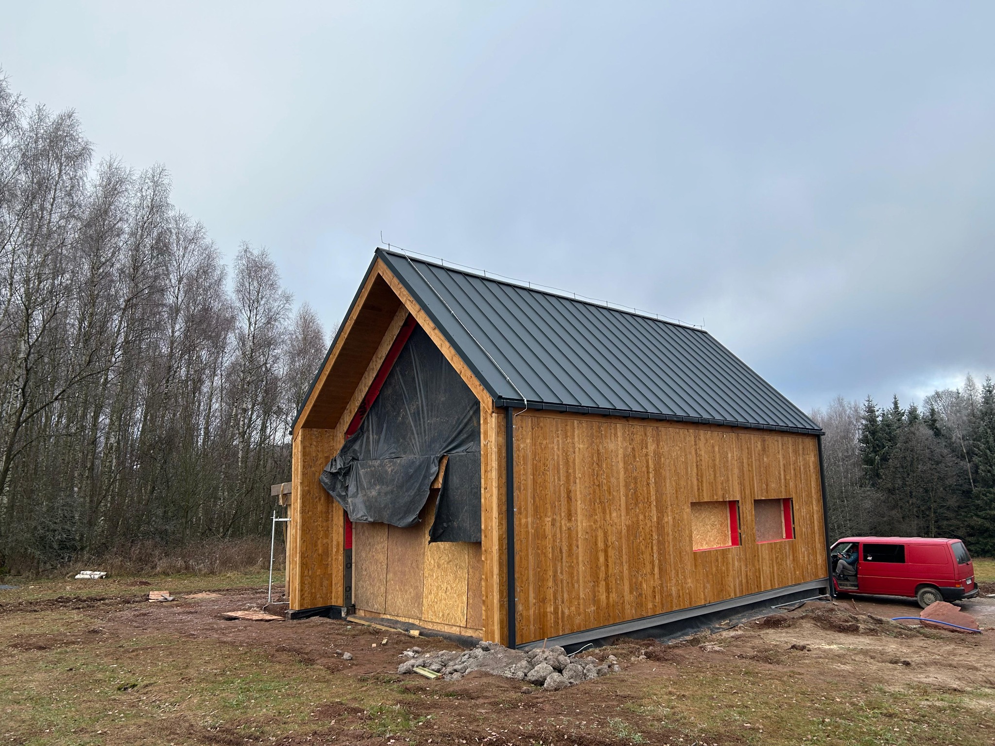 Wznoszony dom szkieletowy z drewnianą elewacją i czarnym dachem na tle lasu. Część ściany zakryta folią. Czerwony bus zaparkowany obok.
