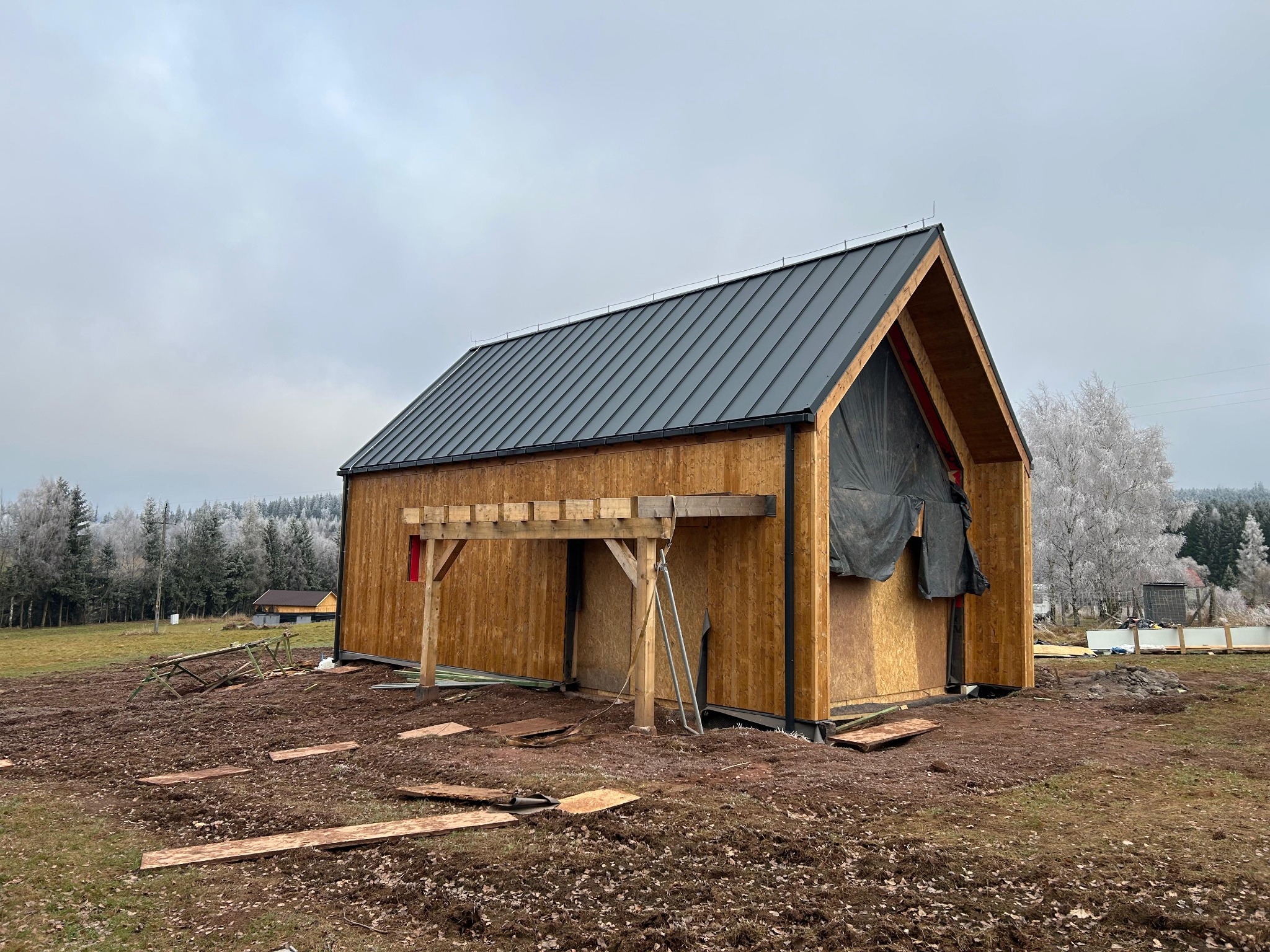 Surowy, drewniany dom szkieletowy z ciemnym dachem na tle zimowego krajobrazu. Widoczne elementy konstrukcyjne i przygotowania do dalszych prac wykończeniowych.