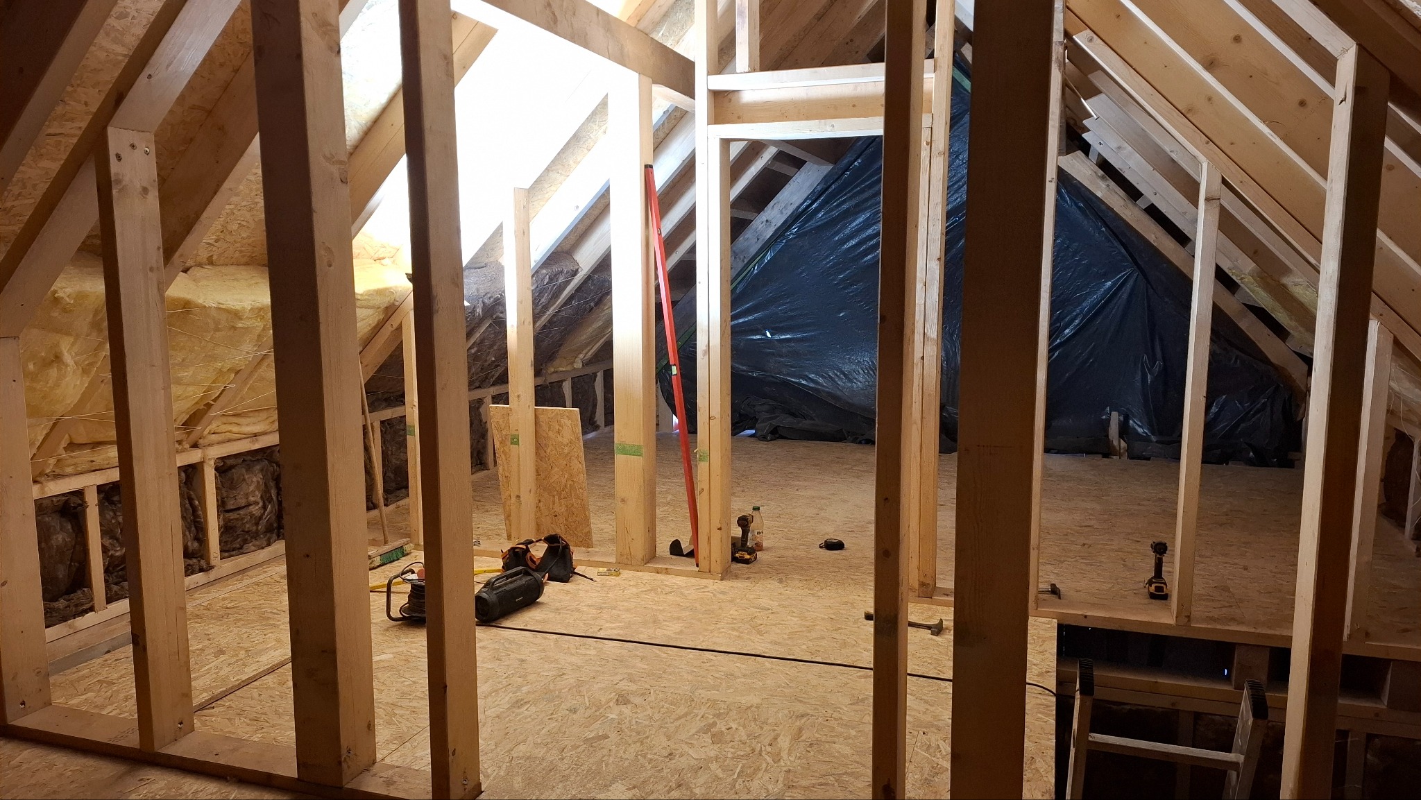 Szkielet drewniany poddasza z widoczną izolacją, narzędziami i folią ochronną. Ujęcie z poziomu podłogi, ukazujące konstrukcję w trakcie budowy.