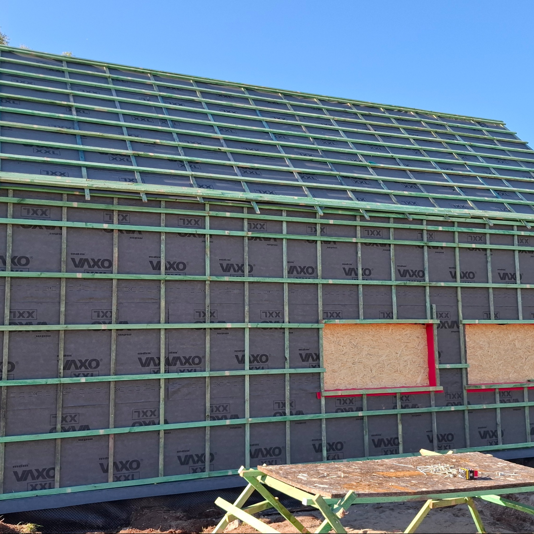 Konstrukcja szkieletowa domu z membraną VAXO XXL, widoczne drewniane elementy i fragmenty okien zabite płytą OSB. Na pierwszym planie stół roboczy z narzędziami.